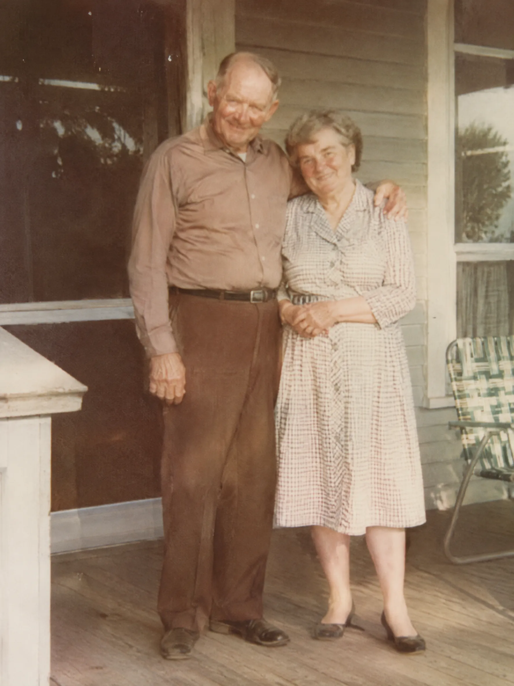 A faded 1960s family photograph of an elderly couple smiling on a wooden porch, soft film grain, warm nostalgic tones