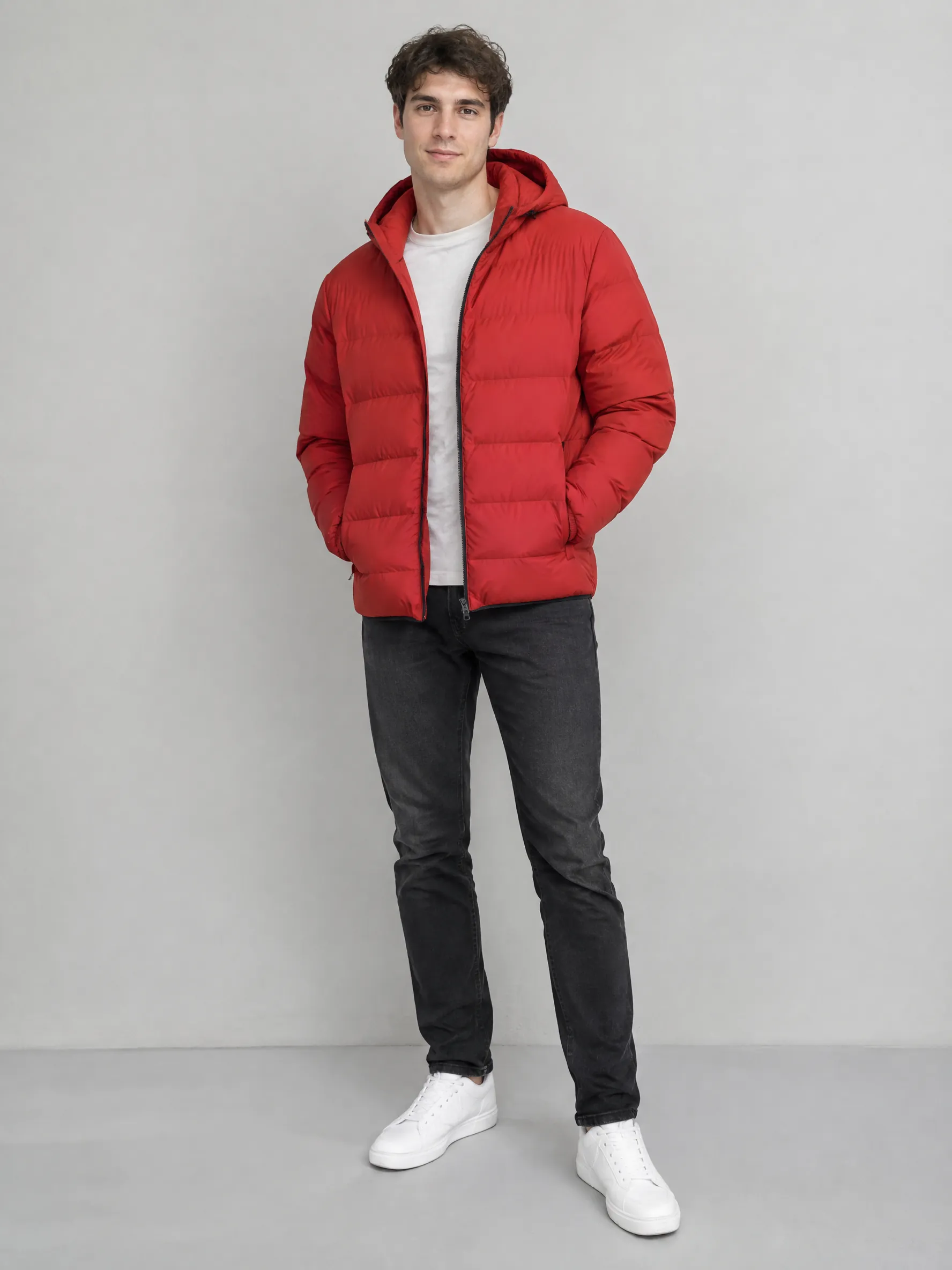 Full-body photo of a young man in a red puffer jacket and white sneakers standing against a plain light grey wall, soft studio light